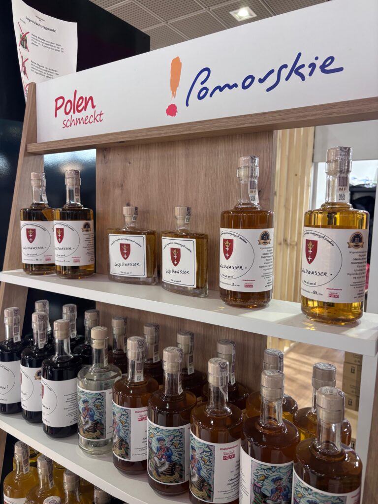 Regał z napisem POLEN SCHMECKT i logo POMORSKIE. na półkach stoją butelki z trunkiem wysokoprocentowym. Na etykietach trunków herb Gdańska
